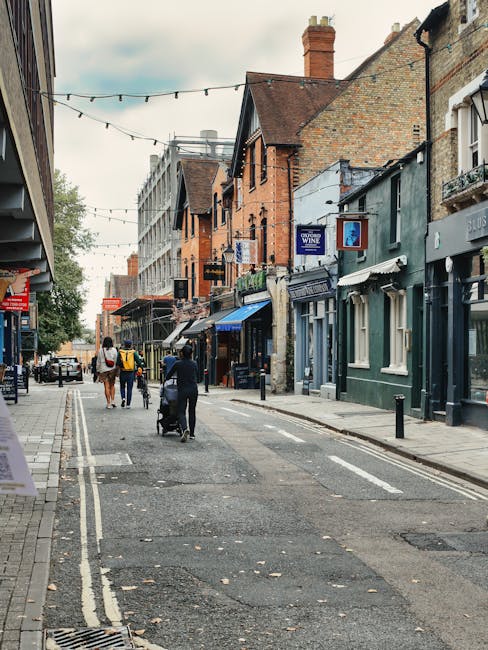 A street scene in Old Coulsdon showing a narrow urban road lined with mixed-use buildings, including shops and residences with brick and painted facades. Pedestrians, including a person pushing a stroller and others walking, are visible along the pavement, which is partially obstructed by bollards. Overhead, string lights are hung across the street, and a few parked cars can be seen in the distance. The buildings feature a variety of architectural styles, with some having signage related to local businesses such as a wine shop and other retail outlets. The scene suggests a typical neighbourhood street where home and commercial activities occur, relevant to house removals and moving logistics, as exemplified by the context of [COMPANY_NAME] providing removals services in the area.