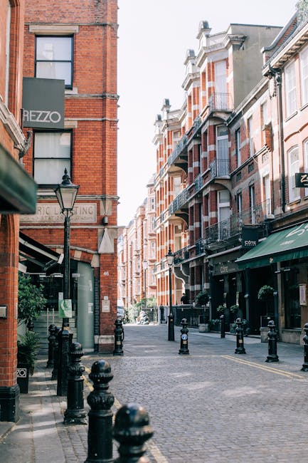 A narrow urban street scene in Old Coulsdon featuring red-brick Victorian-style buildings with bay windows and balconies, some with decorative wrought iron railings. The street is paved with cobblestones and lined with black bollards along the curb, with a few street lamps and greenery in planters. To the left, there is a signage for 'HEZZO' and a shop window with a green awning, while further down the street, the facades of residential and commercial properties are visible, some with white window frames and brick detailing. The scene appears to be daytime with bright, natural lighting, and no visible activity or vehicles. This environment is typical of a quiet residential and shopping corridor suitable for house removals or furniture transport when preparing for a home relocation, with [COMPANY_NAME] providing professional moving services evident on the site.