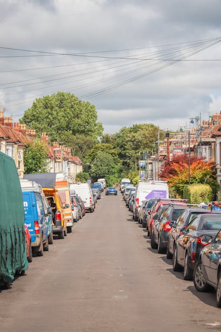 A street scene in Old Coulsdon showing a narrow urban road lined with mixed-use buildings, including shops and residences with brick and painted facades. Pedestrians, including a person pushing a stroller and others walking, are visible along the pavement, which is partially obstructed by bollards. Overhead, string lights are hung across the street, and a few parked cars can be seen in the distance. The buildings feature a variety of architectural styles, with some having signage related to local businesses such as a wine shop and other retail outlets. The scene suggests a typical neighbourhood street where home and commercial activities occur, relevant to house removals and moving logistics, as exemplified by the context of [COMPANY_NAME] providing removals services in the area.