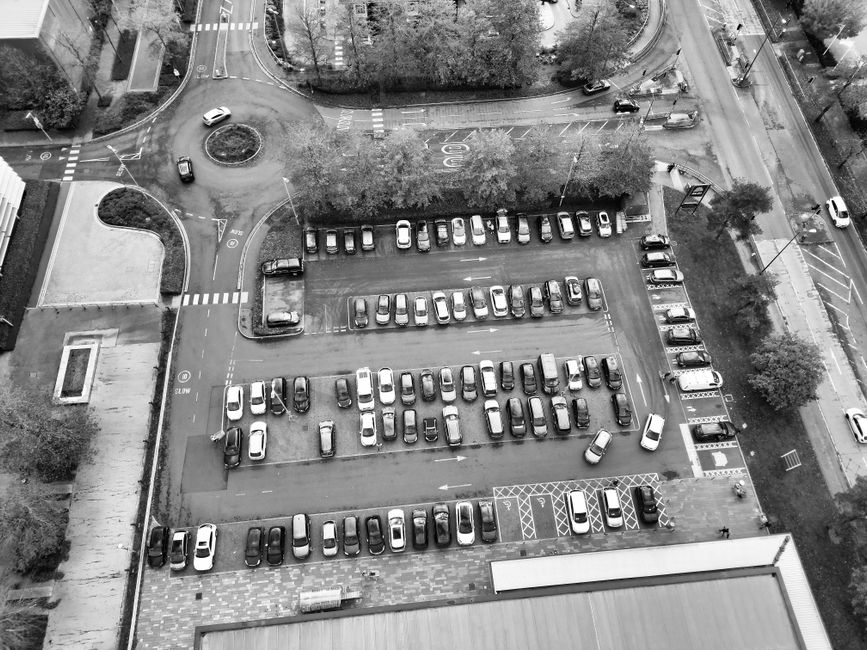 An aerial view of a parking lot situated beside a residential area with multiple lanes for vehicle movement. The lot contains numerous parked cars arranged in parallel rows, with some facing different directions. Surrounding the parking area are trees, a small landscaped section, and nearby roads with clear traffic markings, including pedestrian crossings and turning lanes. On the ground near the entrance to the parking lot, several cardboard moving boxes, wrapped furniture pieces, and packing materials are visible, indicating a home relocation process. A man with a van, part of the Man with Van Old Coulsdon removals service, can be seen actively loading or unloading furniture and boxes onto or from the vehicle, which is positioned adjacent to the parking area. The scene is set in daylight with natural lighting, and the overall environment reflects a typical urban streetscape supporting logistical considerations like parking permits for Old Coulsdon moves, as outlined in the Croydon Council regulations.