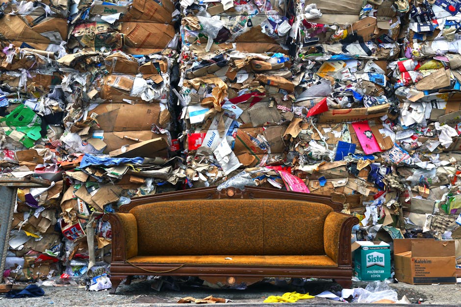 A vintage brown upholstered sofa with wooden frame and intricate carvings on the backrest is positioned outdoors against a large pile of bulky waste materials, including crumpled cardboard boxes, torn paper, and assorted packaging debris, some of which are in plastic wrapping. The waste is stacked high, filling the background and partially covering the pavement area. Nearby, there are cardboard boxes labeled with product names and a small yellow object on the ground. The setting suggests an outdoor space possibly connected to a house or commercial premises involved in waste disposal or clearance, with natural daylight illuminating the scene. This image reflects the type of waste management and furniture removal tasks handled by companies like Man with Van Old Coulsdon during home relocations or bulk waste collection nearby.