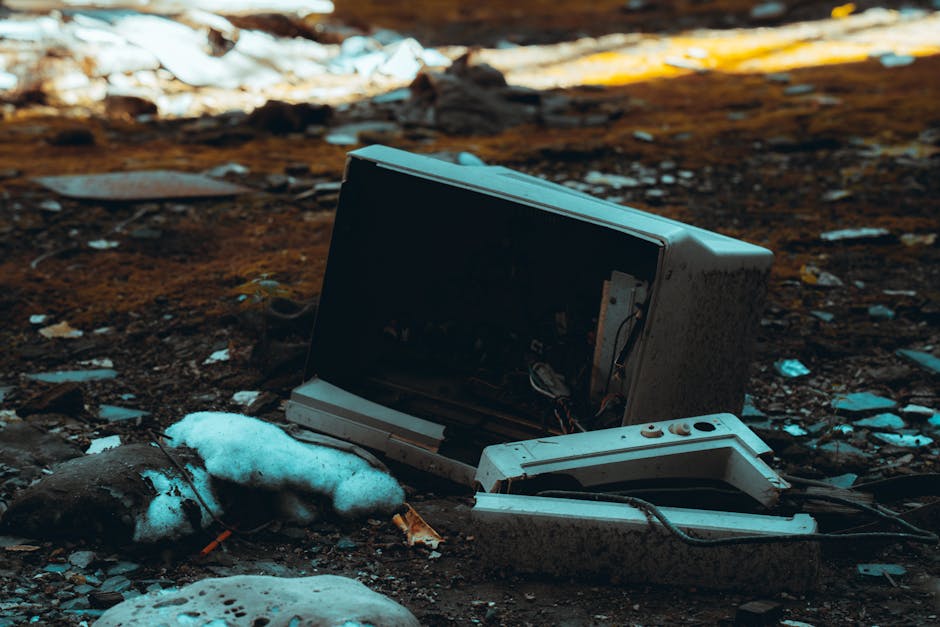 A discarded, broken computer monitor and a detached keyboard lying on the ground outdoors, surrounded by patches of melting snow and muddy soil. The monitor is turned slightly to the side, exposing its internal components and wiring, indicating it is no longer functional. The scene captures the debris and waste materials often associated with bulky waste disposal in residential areas. In the background, there are scattered pieces of paper and other refuse. The environment appears to be a driveway or backyard area, with natural lighting highlighting the contrast between the electronic waste and the surrounding natural elements. This image illustrates the type of bulky waste items that may be involved in home relocation or clearance services provided by companies such as Man with Van Old Coulsdon, highlighting the importance of proper removal and disposal of electronic waste during house moves and waste management processes.
