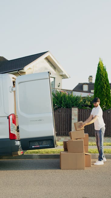 A man wearing a white T-shirt and jeans is standing next to a white van with its rear doors open, loading cardboard boxes onto the pavement outside a residential property. The boxes are of various sizes, some stacked on top of each other, and appear to contain household belongings for a home relocation or furniture transport. The van is positioned close to a driveway, with a suburban street in the background, including a house with a pitched roof, a wood fence, and greenery such as trees and bushes. The scene is illuminated by natural daylight, and the man is in the process of organizing or carrying items as part of packing and moving services. This image exemplifies the loading process typical of professional removals or last-minute move solutions offered by companies like Man with Van Old Coulsdon.