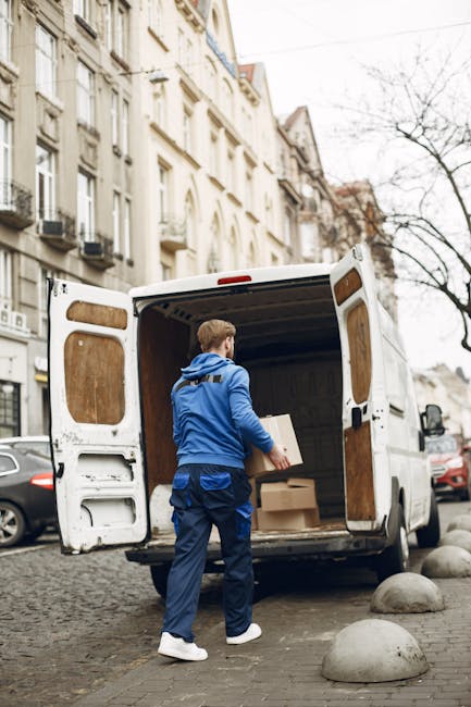 A man wearing a blue jacket, white sneakers, and dark blue trousers is loading cardboard boxes into the open rear of a white moving van parked on a cobblestone street in Old Coulsdon. The van's interior is lined with wooden panels, and several boxes are stacked inside, ready for a home relocation or furniture transport. The background features a row of historic multi-storey buildings with cream facades, bay windows, and black wrought iron balconies, characteristic of the area. Leafless trees are visible along the street, indicating a colder season. Large, smooth concrete bollards separate the parking area from the sidewalk, and other vehicles are parked nearby. The scene captures the process of packing and loading during a house move, with natural daylight illuminating the environment. Man with Van Old Coulsdon provides removals services, including loading and transportation, as exemplified by this activity.