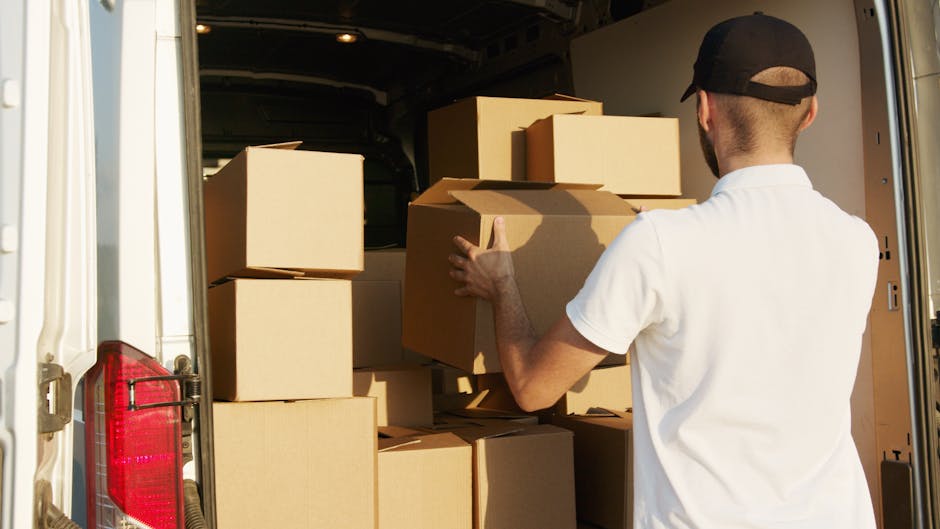 A man wearing a white T-shirt and jeans is standing next to a white van with its rear doors open, loading cardboard boxes onto the pavement outside a residential property. The boxes are of various sizes, some stacked on top of each other, and appear to contain household belongings for a home relocation or furniture transport. The van is positioned close to a driveway, with a suburban street in the background, including a house with a pitched roof, a wood fence, and greenery such as trees and bushes. The scene is illuminated by natural daylight, and the man is in the process of organizing or carrying items as part of packing and moving services. This image exemplifies the loading process typical of professional removals or last-minute move solutions offered by companies like Man with Van Old Coulsdon.