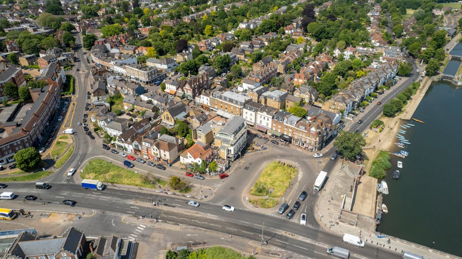 Aerial view of Old Coulsdon High Street, showing a mix of residential and commercial buildings, with narrow pavement spaces and a small parking area adjacent to the street. The image captures vehicles including cars, vans, and a delivery truck parked along the roadside and within a small courtyard, which contains a few trees and patches of grass. To the right, a body of water with moored boats is visible, with a promenade running alongside. The scene includes street lamps, traffic signs, and buildings with brick and tiled facades, illustrating a typical town centre environment suitable for house and furniture removals. The lighting indicates daytime, with clear skies casting shadows across the scene, highlighting the tight urban layout. This setting is relevant for ongoing home relocation and furniture transport activities, supported by the professional removals services of Man with Van Old Coulsdon, particularly during the loading process of items moving in or out of nearby properties.