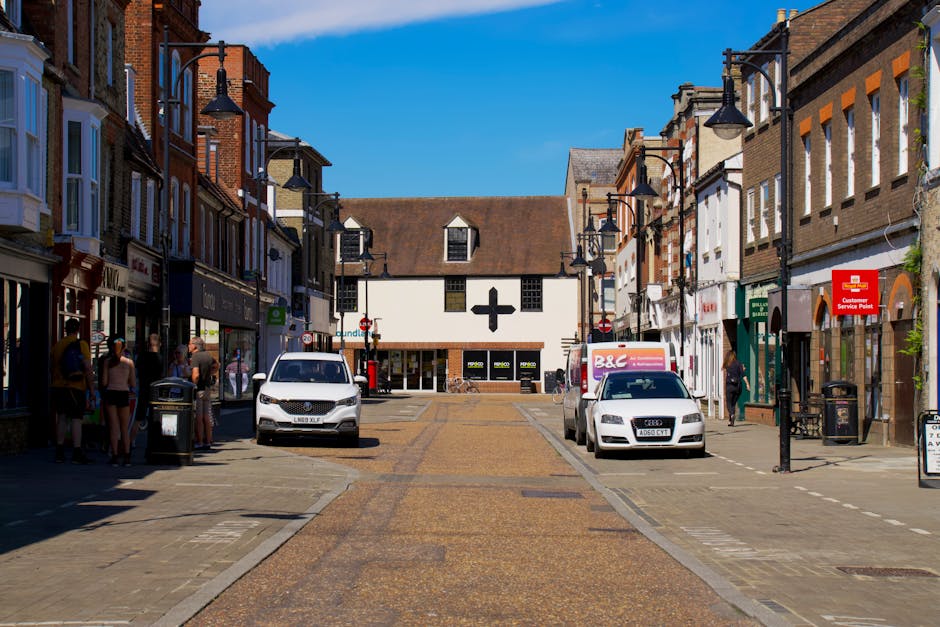 A wide street view in Old Coulsdon High Street showing several small retail shops and businesses housed in traditional brick and weatherboard buildings with large display windows. On the left side of the street, a group of people are gathered, some standing near a black lamp post and a trash bin, suggesting foot traffic typical of a shopping area. Parked along the curb are two white and one silver cars, including an Audi, and a delivery van with branding partially visible. The pavement features a textured, reddish-brown surface, while the roadway is paved with smooth asphalt. Street lamps with black metal fixtures line both sides of the street, and a prominent white building with a black cross on its facade is visible at the end of the street, possibly a pharmacy or medical centre. The scene is well-lit with clear blue sky overhead, and the overall environment indicates a busy commercial area suitable for local shopping, with potential for home relocation and furniture transport services, as coordinated by local removals specialists like Man with Van Old Coulsdon.