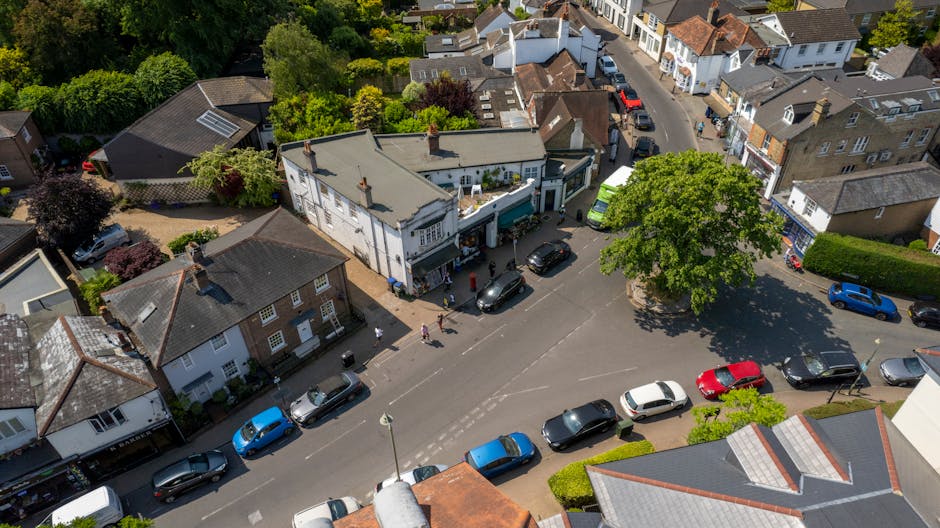 Aerial view of Old Coulsdon High Street, showing a mix of residential and commercial buildings, with narrow pavement spaces and a small parking area adjacent to the street. The image captures vehicles including cars, vans, and a delivery truck parked along the roadside and within a small courtyard, which contains a few trees and patches of grass. To the right, a body of water with moored boats is visible, with a promenade running alongside. The scene includes street lamps, traffic signs, and buildings with brick and tiled facades, illustrating a typical town centre environment suitable for house and furniture removals. The lighting indicates daytime, with clear skies casting shadows across the scene, highlighting the tight urban layout. This setting is relevant for ongoing home relocation and furniture transport activities, supported by the professional removals services of Man with Van Old Coulsdon, particularly during the loading process of items moving in or out of nearby properties.