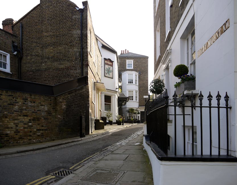 A narrow residential street in Old Coulsdon with cobbled pavement and a slight incline, featuring tightly packed buildings on either side. On the right, a white building with a black wrought iron fenc