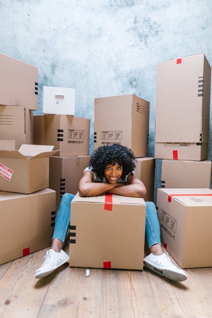 A young woman with curly black hair, wearing a white t-shirt, blue jeans, and white sneakers, is sitting on a light wooden floor surrounded by numerous cardboard boxes of various sizes, some sealed with red packing tape and others partially open. She is smiling and resting her head on her crossed arms on top of one of the boxes, indicating a relaxed moment during a home relocation or packing process. The background features a plain light blue wall with a slightly textured finish, and the boxes are stacked against it. The scene appears to be indoors, near the doorway or main interior space of a property, with the boxes ready for transportation or storage. This image illustrates the packing and moving activities typical of services provided by Man with Van Old Coulsdon, fitting into the context of furniture transport and home relocation preparations as part of professional removals or moving services.
