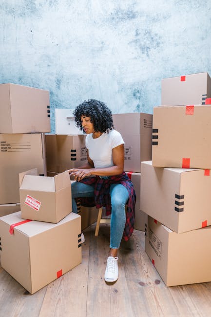 A woman with curly dark hair, dressed in a white t-shirt and blue jeans, is seated on a wooden chair inside a room with a light blue textured wall. She is surrounded by numerous cardboard boxes of various sizes, some sealed with red and black tape, and others open with flaps folded back. The boxes are placed on the wooden floor and stacked against the wall, indicating the process of packing for a house relocation. The woman appears to be organizing or sealing a box, with additional boxes around her suggesting preparations for moving house. A plaid shirt is tied around her waist. This scene is part of a home moving or packing process, associated with professional removal services, such as those provided by Man with Van Old Coulsdon, and reflects typical packing and furniture transport activities involved in moving logistics near Coulsdon South station.