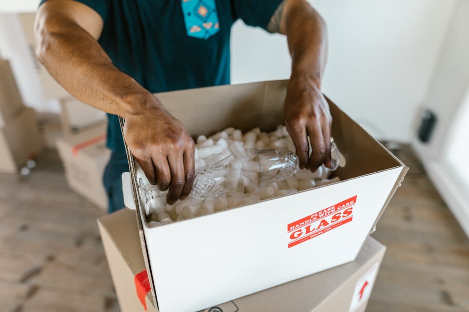 A young woman with curly black hair, wearing a white t-shirt, blue jeans, and white sneakers, is sitting on a light wooden floor surrounded by numerous cardboard boxes of various sizes, some sealed with red packing tape and others partially open. She is smiling and resting her head on her crossed arms on top of one of the boxes, indicating a relaxed moment during a home relocation or packing process. The background features a plain light blue wall with a slightly textured finish, and the boxes are stacked against it. The scene appears to be indoors, near the doorway or main interior space of a property, with the boxes ready for transportation or storage. This image illustrates the packing and moving activities typical of services provided by Man with Van Old Coulsdon, fitting into the context of furniture transport and home relocation preparations as part of professional removals or moving services.