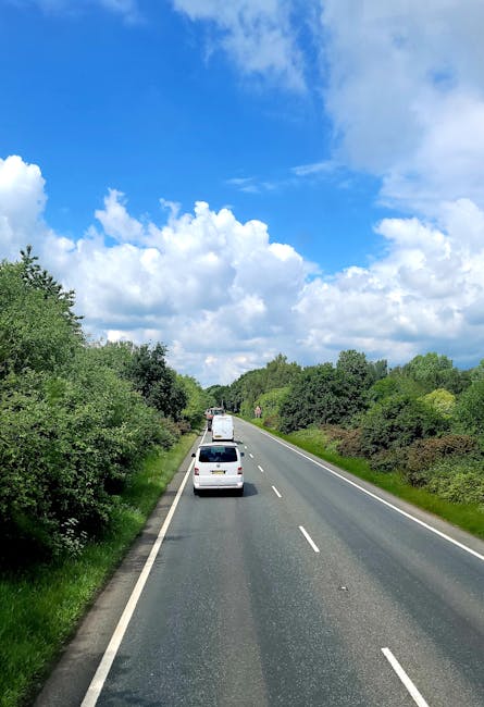 A two-lane rural road with a slight incline, lined with dense green foliage on both sides, leading into a distance under a clear blue sky filled with large, fluffy white clouds. Several vehicles, incl