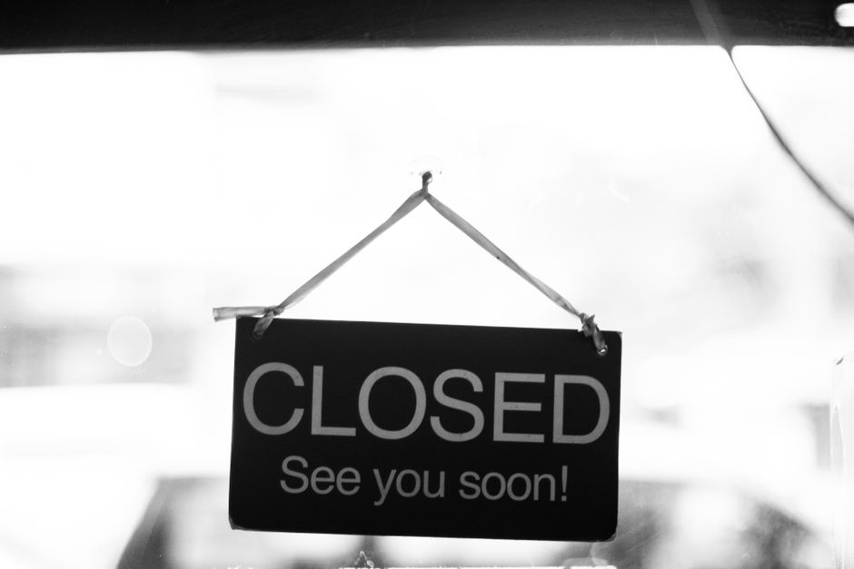 Black and white photograph of a rectangular 'CLOSED' sign with the message 'See you soon!' hanging on a door or window during daytime. The sign is suspended by a white string attached to the top corners, with metal eyelets reinforcing attachment points. The background is out of focus, suggesting an outdoor street environment, possibly near a property involved in a house relocation or removal process. The lighting indicates natural daylight, with a bright overcast sky reflected on the glass surface. The image may be used to illustrate interruptions or temporary closures related to house removals, parking restrictions, or street permits for moving services such as those offered by Man with Van Old Coulsdon.
