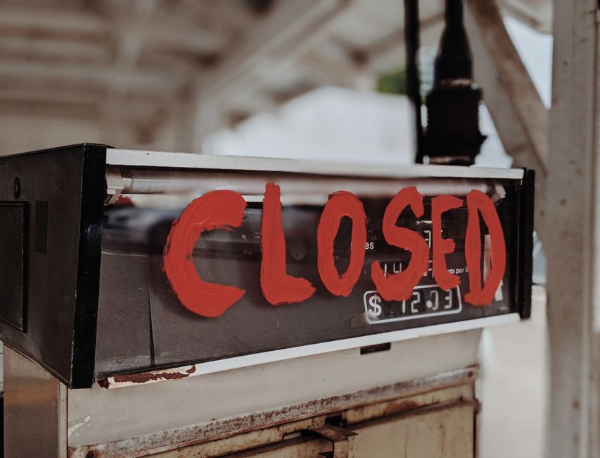 A close-up view of a vintage cash register situated inside a property, with a red, hand-painted sign reading 'CLOSED' placed prominently on its front. The cash register has a black exterior with a visible coin compartment and a metallic handle, and it rests on a wooden surface that shows signs of wear. In the background, part of a wooden structure and some blurred objects can be seen, indicating an indoor environment, possibly a storeroom or a part of a house awaiting a home relocation or packing process. The setting is well-lit with natural light, highlighting the details of the cash register and the painted sign, conveying a scene related to house removals or packing within a property managed by Man with Van Old Coulsdon.