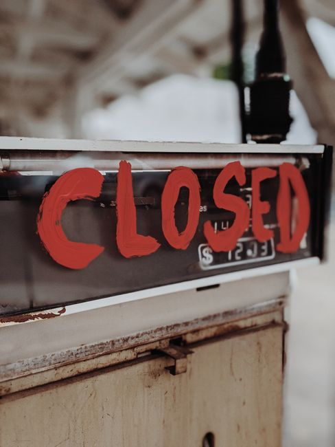 Black and white photograph of a rectangular 'CLOSED' sign with the message 'See you soon!' hanging on a door or window during daytime. The sign is suspended by a white string attached to the top corners, with metal eyelets reinforcing attachment points. The background is out of focus, suggesting an outdoor street environment, possibly near a property involved in a house relocation or removal process. The lighting indicates natural daylight, with a bright overcast sky reflected on the glass surface. The image may be used to illustrate interruptions or temporary closures related to house removals, parking restrictions, or street permits for moving services such as those offered by Man with Van Old Coulsdon.