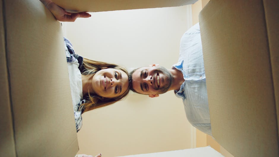 A man and a woman are lying inside cardboard moving boxes in an indoor setting, looking up towards the camera with smiles. The woman has light brown hair and is wearing a plaid shirt, while the man has short hair, a beard, and is dressed in a light-colored shirt with rolled-up sleeves. They are positioned near the edges of the open boxes, which are placed on either side of them, suggesting a home relocation process. The scene is lit with natural or soft indoor lighting, highlighting the materials of the boxes and the casual atmosphere of packing and moving preparations. This image, associated with house removals services by Man with Van Old Coulsdon, visually represents the packing and loading stage of a household move, emphasizing the careful handling of belongings in furniture transport and load management.