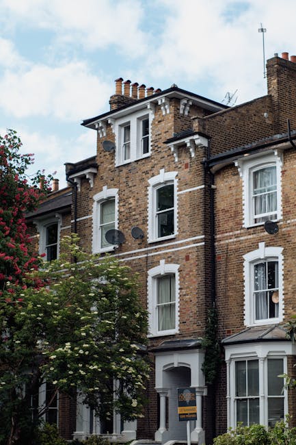 Photograph of a multi-storey brick residential building with white framed sash windows and decorative white brickwork accents along the edges and around the window openings. Several satellite dishes are mounted on the exterior walls, and the roof features multiple chimneys and a weather vane. A lush green tree with flowering branches partially obscures the lower part of the building, set against a partly cloudy sky. This image depicts a typical London-style house, aligning with house removal and moving services offered by Man with Van Old Coulsdon, particularly relevant for clients planning home relocations from Farthing Downs to London homes. The building's exterior presents a classic, period-appropriate façade suitable for interior packing and furniture transport during a home relocation process.