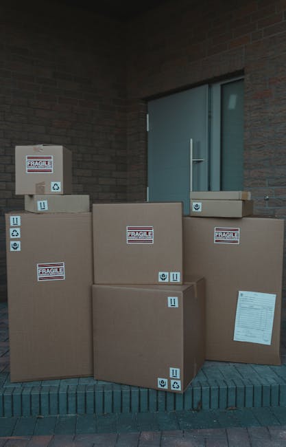 Several large cardboard moving boxes, some labeled 'Fragile' with handling symbols, are stacked outside a building near a grey door, resting on a brick pavement. The boxes vary in size, with some plac